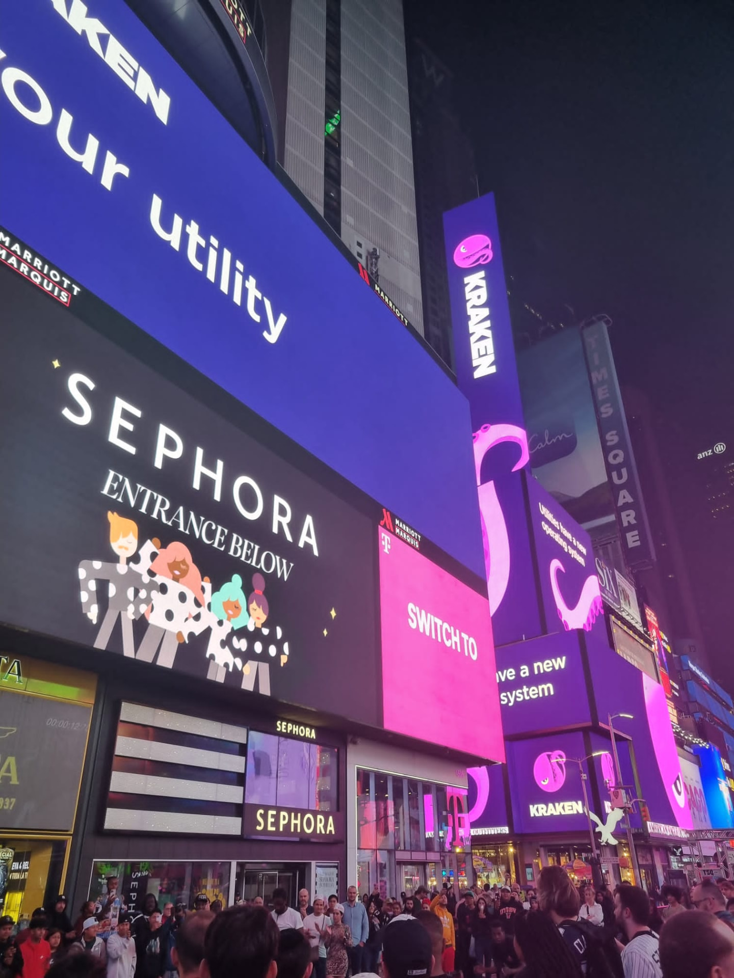 Crowds in Times Square beneath Kraken screens reading 'switch to your utility'